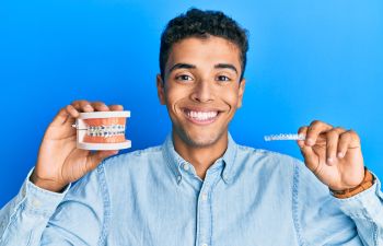 A teenage Afro-American boy holding a clear aligner in one hand and a dental model with metal braces in the other hand.